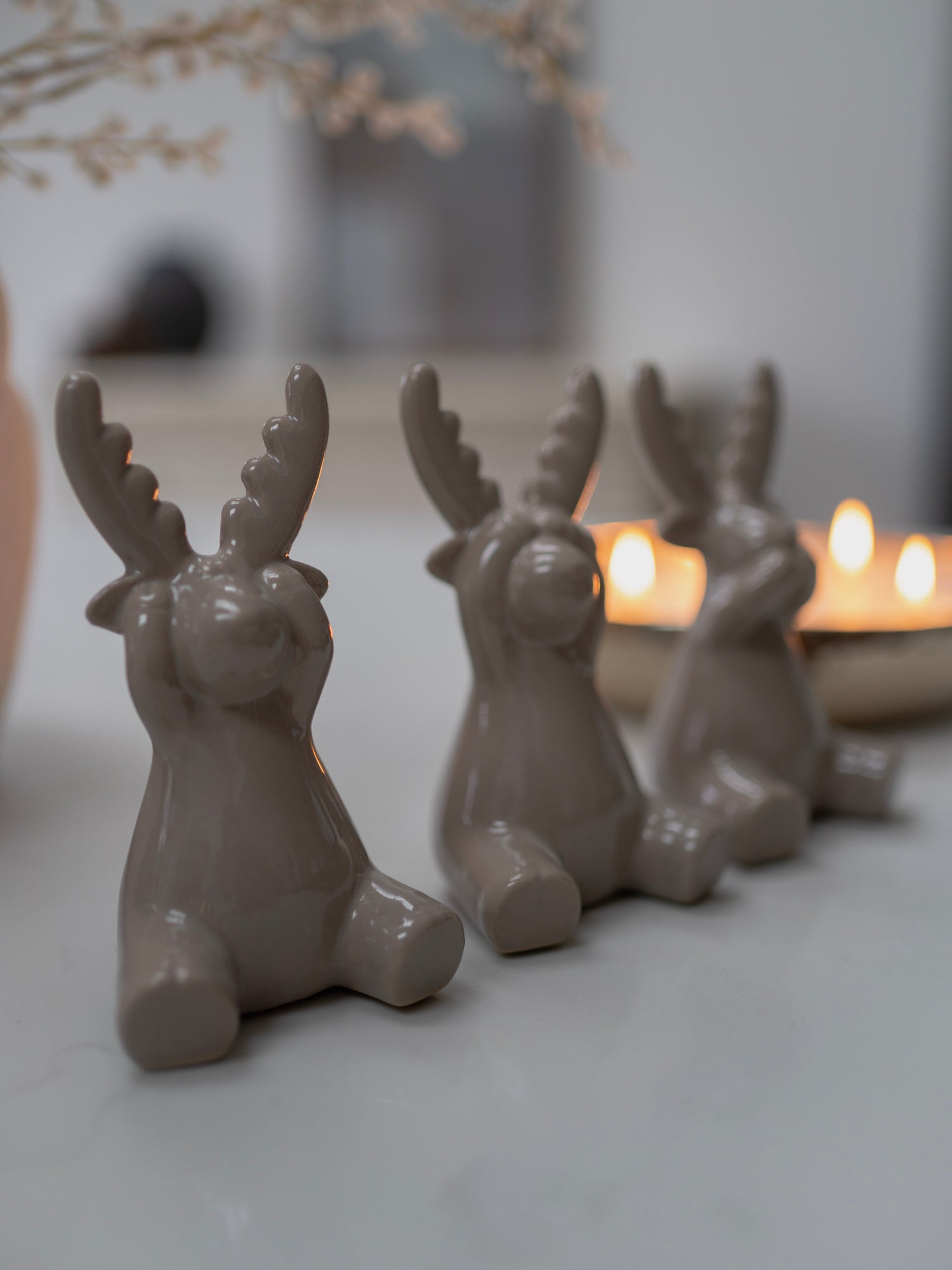Three grey ceramic deer figurines with antlers on a light surface with candles in the background.