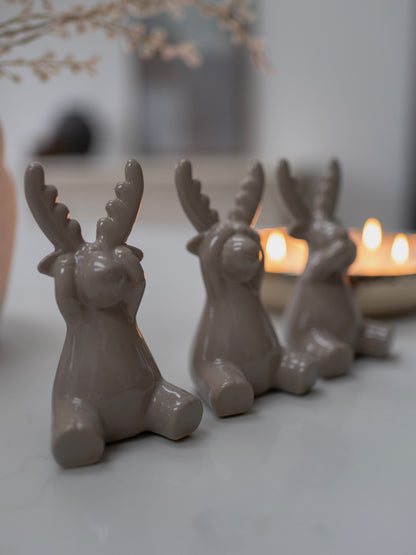 Three grey ceramic deer figurines with antlers on a light surface with candles in the background.