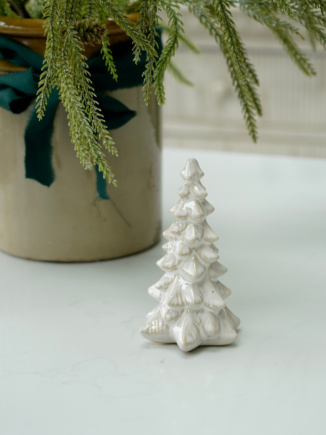White ceramic Christmas tree figurine on a light surface with a plant in the background.