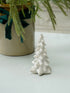 White ceramic Christmas tree figurine on a light surface with a plant in the background.