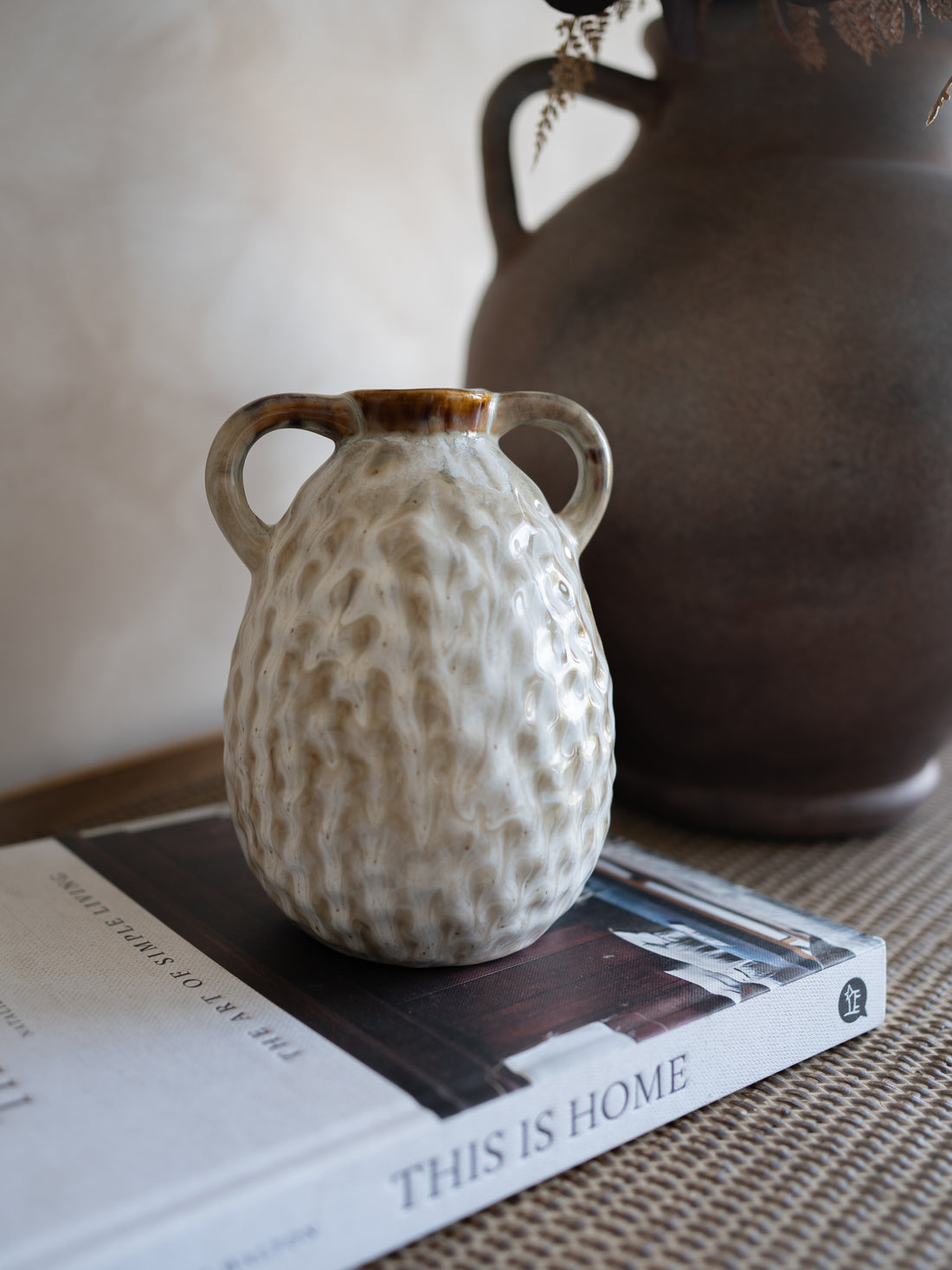 Small textured ceramic vase on a book with a larger brown vase in the background.