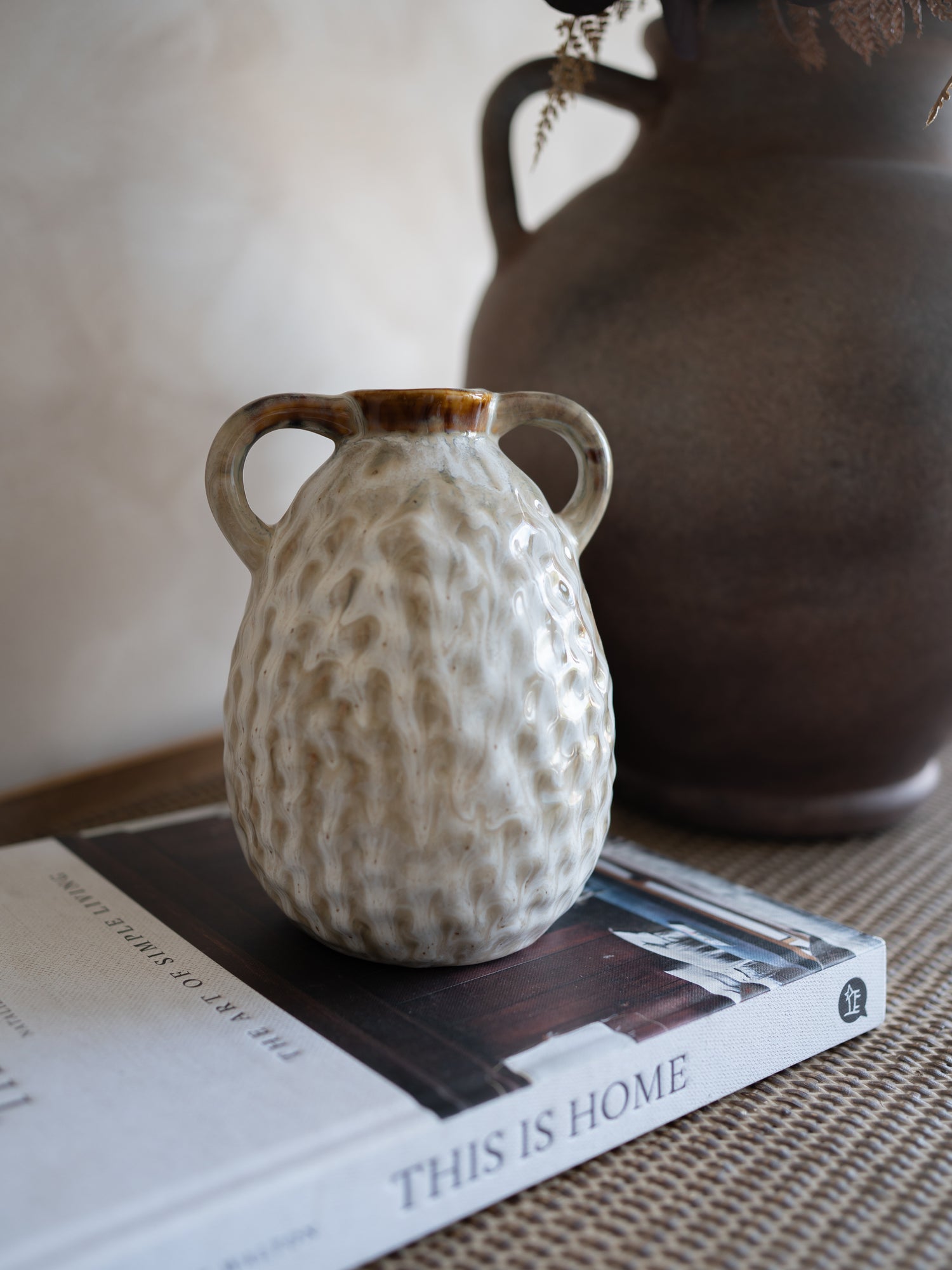 Small textured ceramic vase on a book with a larger brown vase in the background.