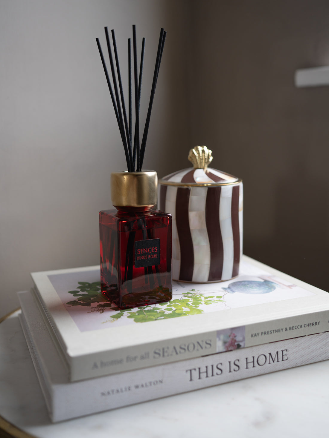 A reed diffuser bottle with a gold cap and black reeds sits on top of two stacked books, alongside a striped decorative container with a gold lid, in a softly lit indoor setting.