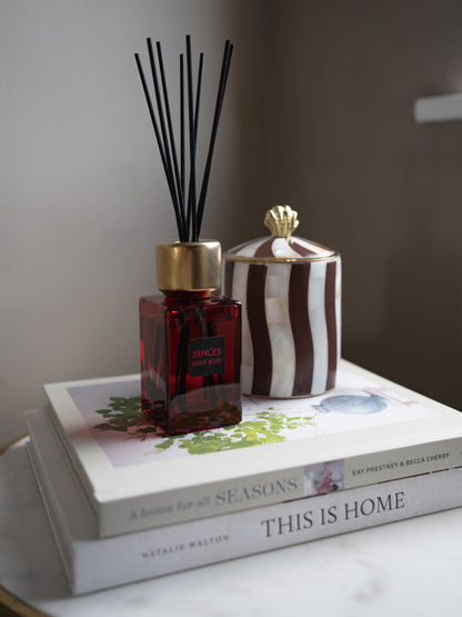 A reed diffuser bottle with a gold cap and black reeds sits on top of two stacked books, alongside a striped decorative container with a gold lid, in a softly lit indoor setting.