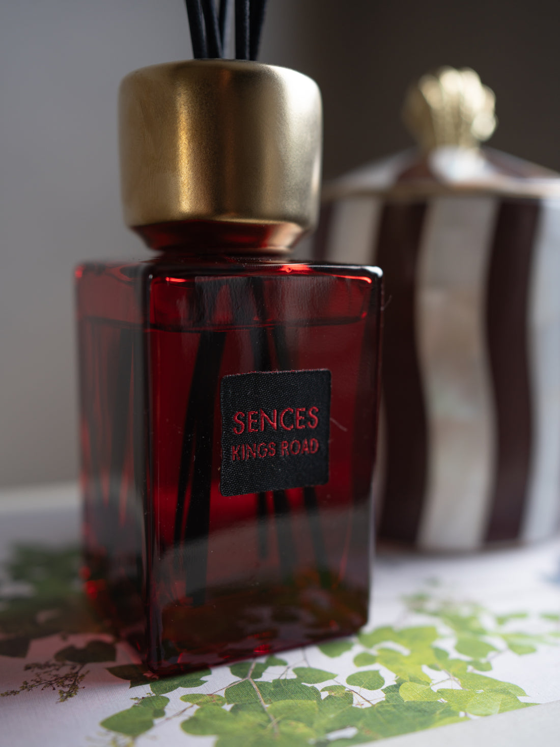 A Scented Kings Road reed diffuser bottle with a deep red glass body, a metallic gold cap, and black reeds, set against a softly blurred background with another decorative bottle.