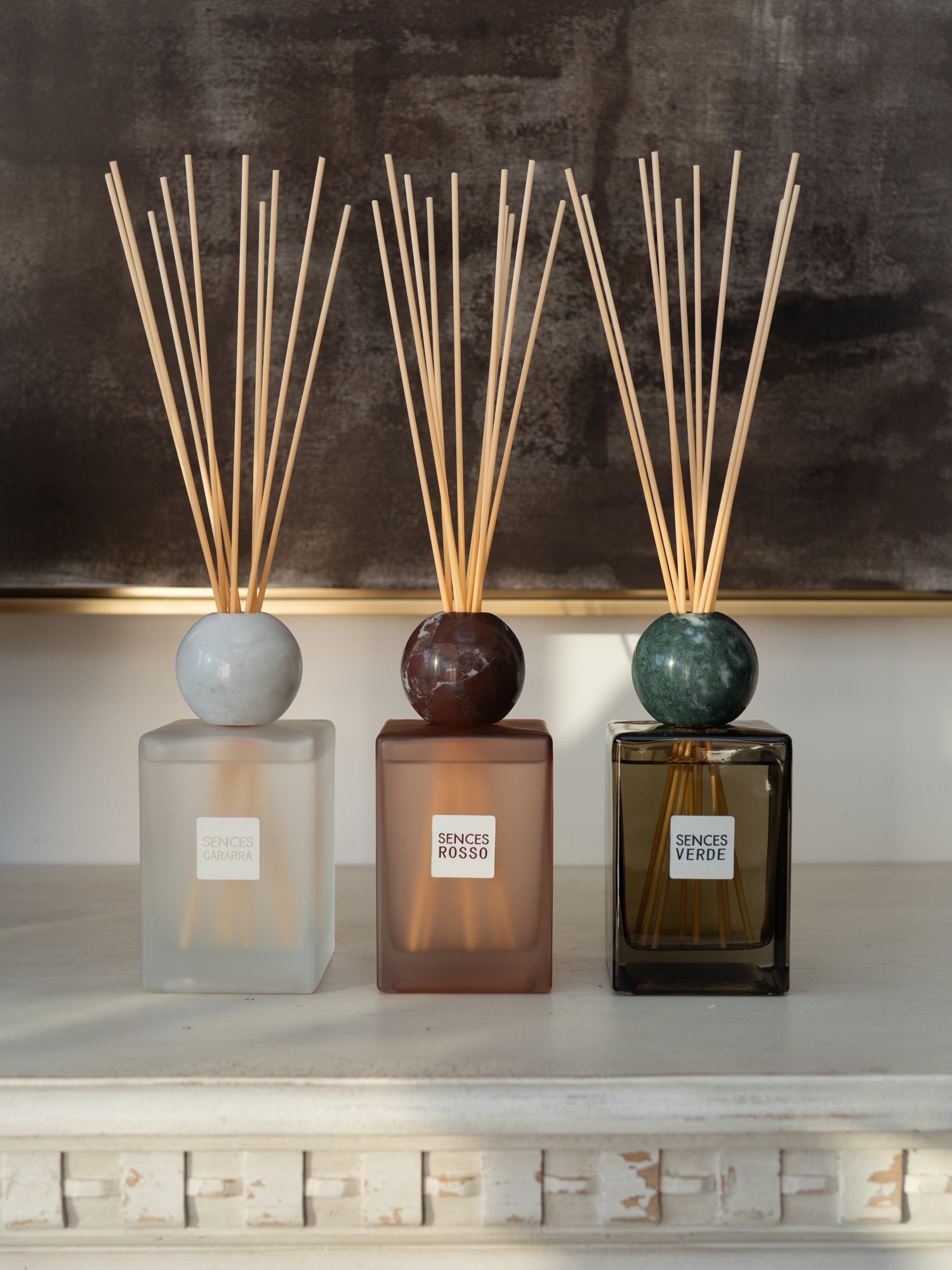 Three reed diffusers with marble-style round tops and natural reeds are arranged on a white mantelpiece against a dark textured wall. The diffusers have frosted, smoky, and dark amber glass bodies with labels reading SENCES ROSSO, SENCES VERDE, and a third label partially visible.