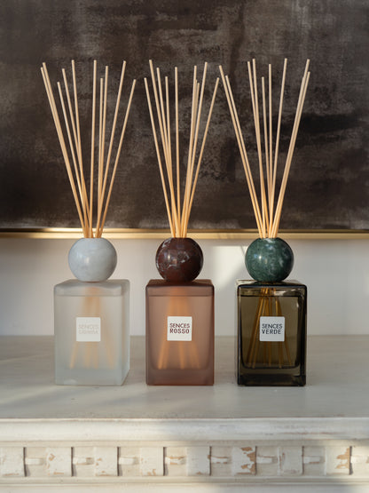 Three reed diffusers with marble-style round tops and natural reeds are arranged on a white mantelpiece against a dark textured wall. The diffusers have frosted, smoky, and dark amber glass bodies with labels reading SENCES ROSSO, SENCES VERDE, and a third label partially visible.