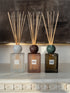 Three reed diffusers with marble-style round tops and natural reeds are arranged on a white mantelpiece against a dark textured wall. The diffusers have frosted, smoky, and dark amber glass bodies with labels reading SENCES ROSSO, SENCES VERDE, and a third label partially visible.