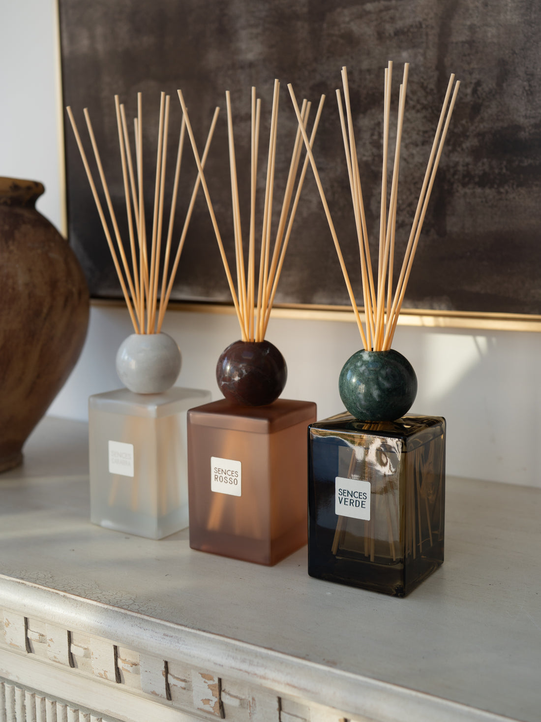 Three reed diffusers with square glass bottles and natural reed sticks are arranged on a light console table. The diffusers have frosted white, amber-brown, and dark smoky green bottles with round tops.