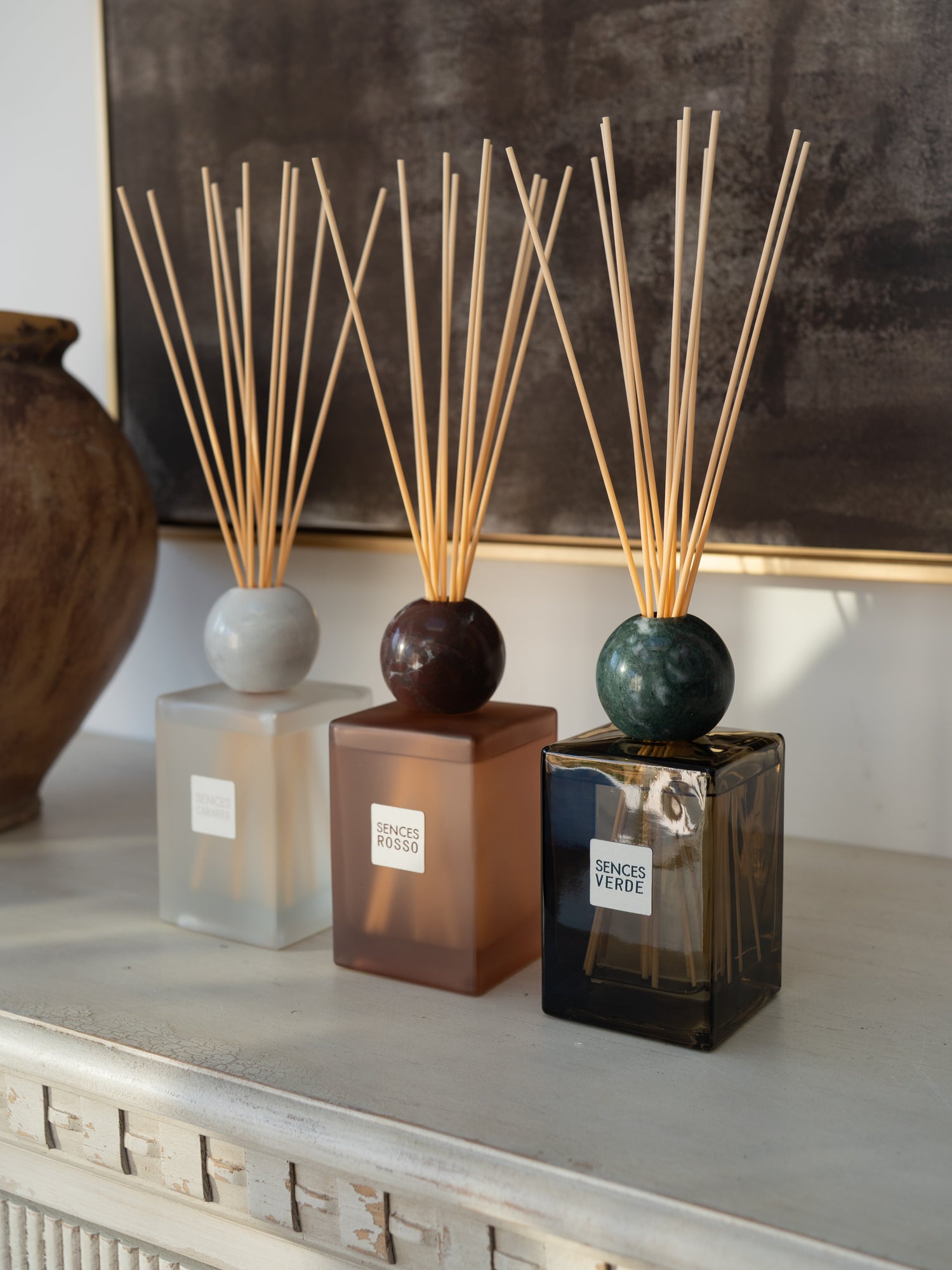 Three reed diffusers with square glass bottles and natural reed sticks are arranged on a light console table. The diffusers have frosted white, amber-brown, and dark smoky green bottles with round tops.