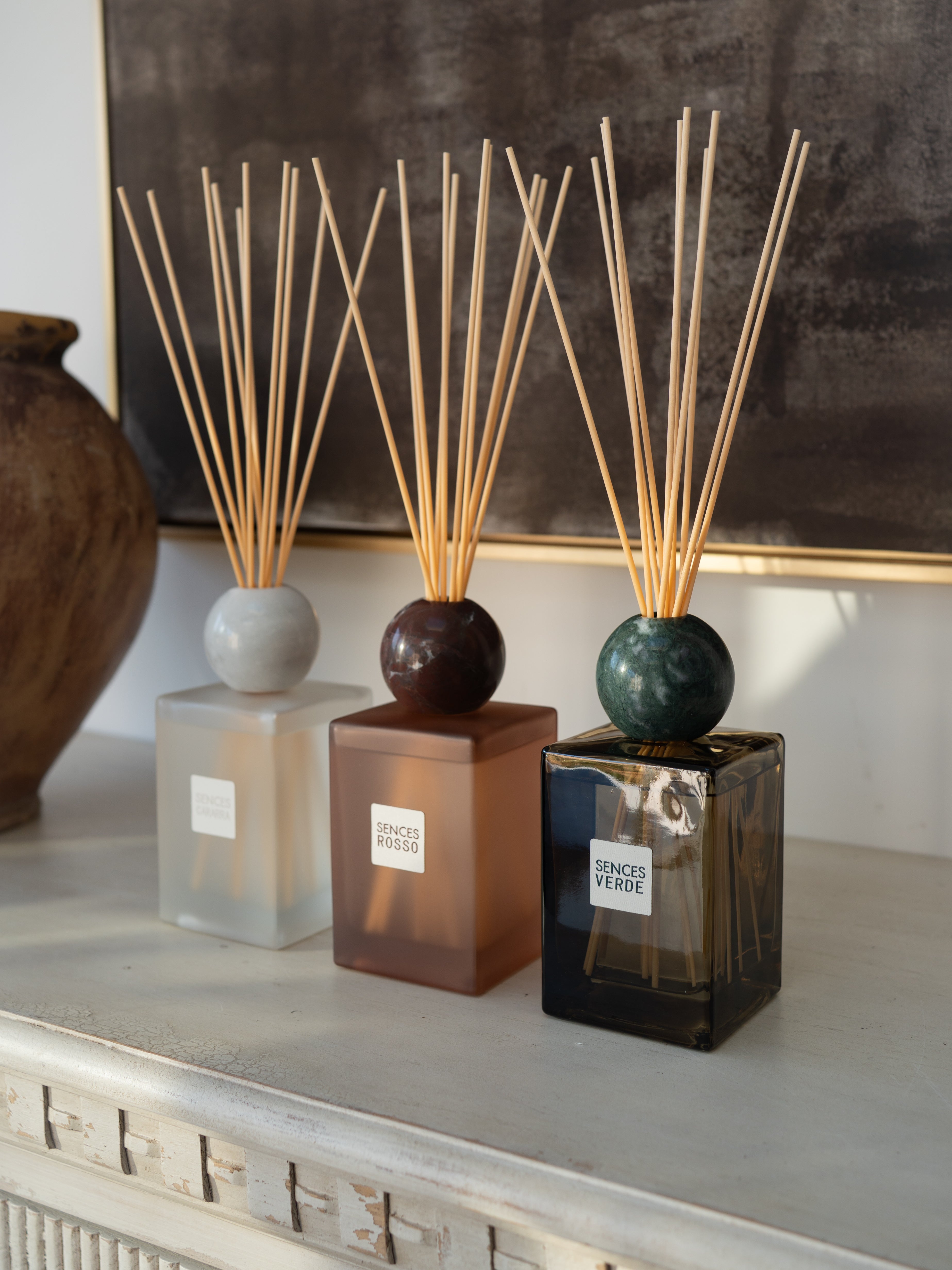 Three reed diffusers with square glass bottles and natural reed sticks are arranged on a light console table. The diffusers have frosted white, amber-brown, and dark smoky green bottles with round tops.