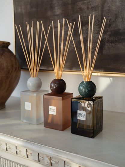 Three reed diffusers with square glass bottles and natural reed sticks are arranged on a light console table. The diffusers have frosted white, amber-brown, and dark smoky green bottles with round tops.