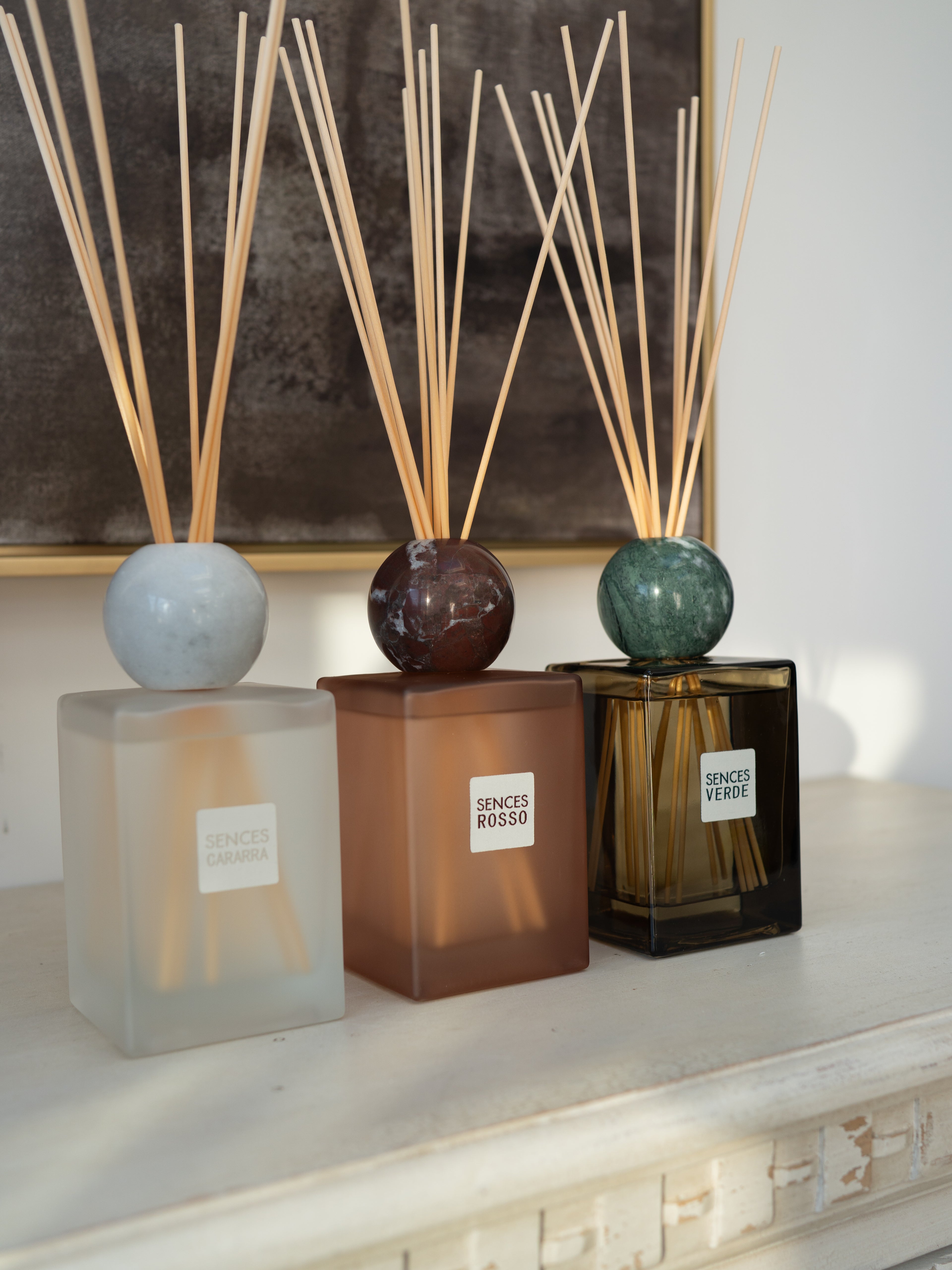 Three reed diffusers with square glass bottles and spherical tops are arranged on a white mantelpiece. The bottles are frosted translucent white, warm amber-brown, and dark amber-brown, each holding natural reeds.