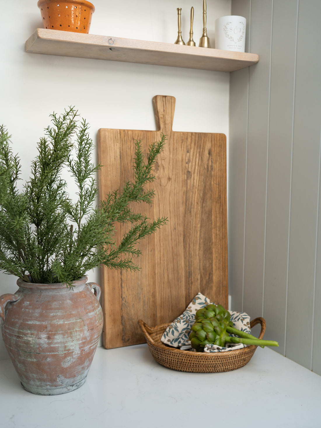 Wooden elm cutting board with a basket of artichokes and a vase of greenery on a white surface.