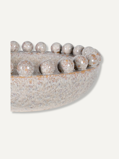A close-up of a decorative ceramic bowl with a textured, marbled finish in beige, gray, and white, featuring a row of small spherical protrusions around the rim, shown on a light off-white background.