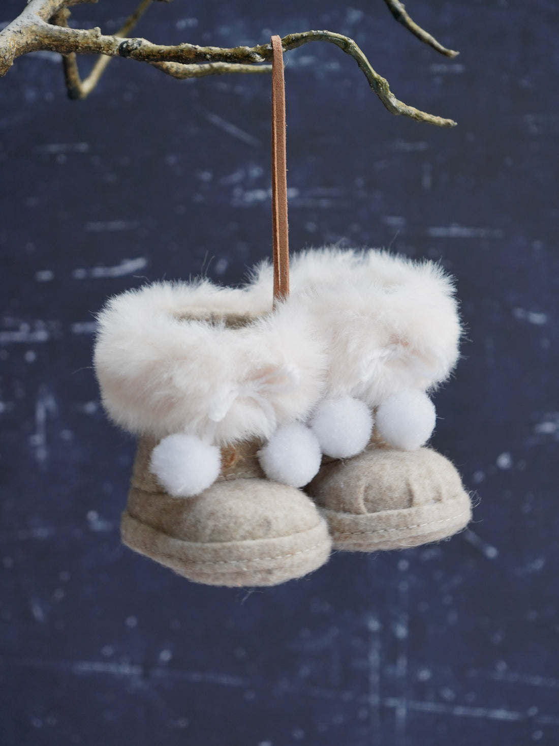 Decorative boot ornaments with white fur and pom-poms hanging from a branch against a dark background.