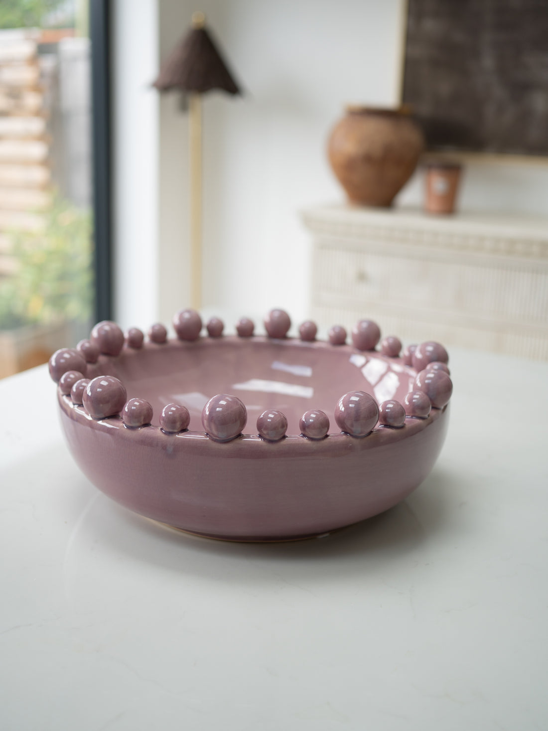 A round, lilac ceramic bowl with a raised beaded rim, set on a light-colored tabletop with a softly blurred indoor background.