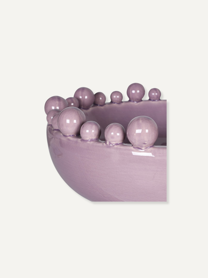 A close-up of a glossy lavender-purple bowl or planter with a row of matching decorative beads along the rim, shown against a plain light beige background.