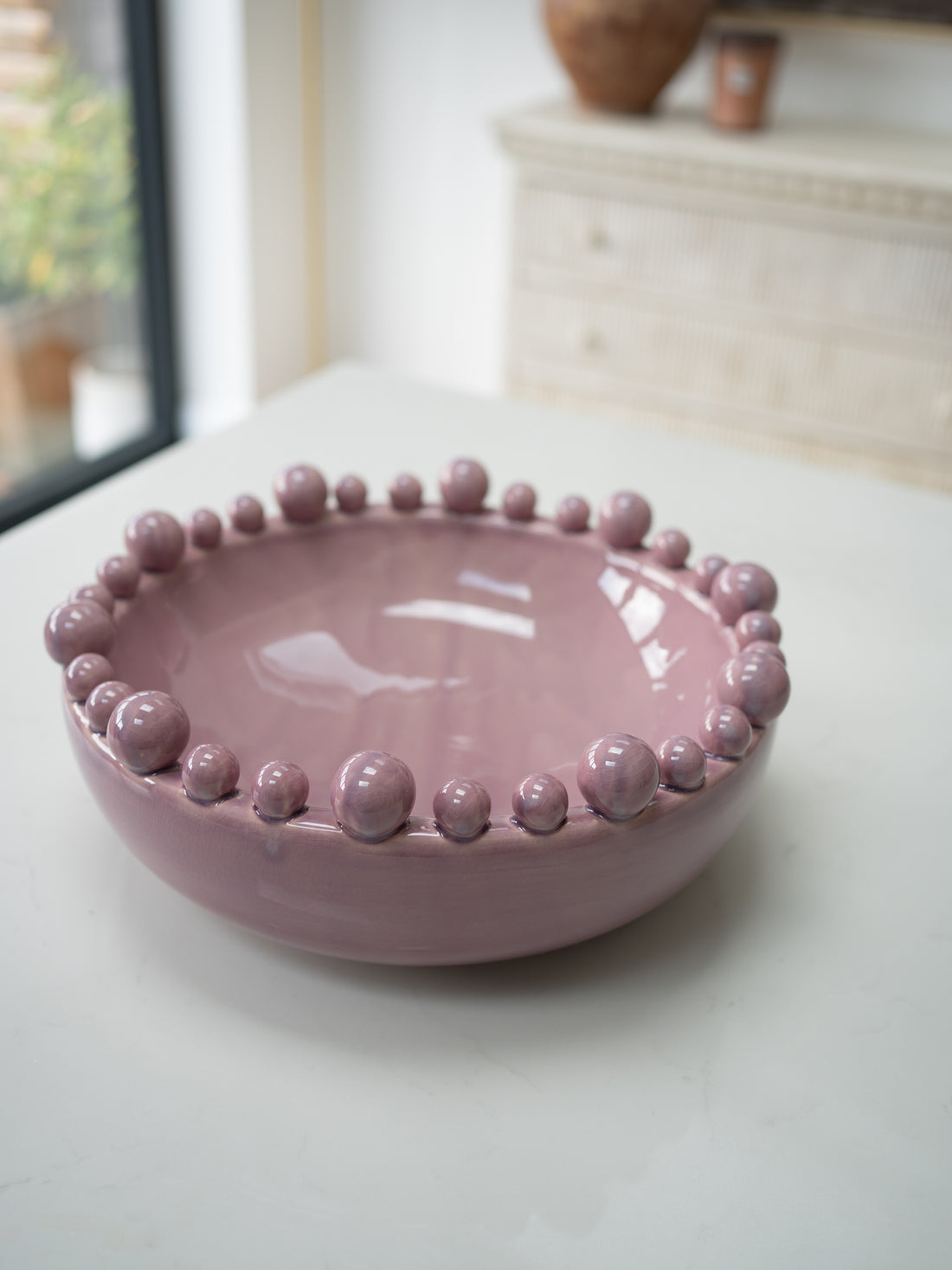 A dusty pink glazed ceramic bowl with a raised beaded rim, set on a light-colored tabletop in a softly blurred indoor setting.