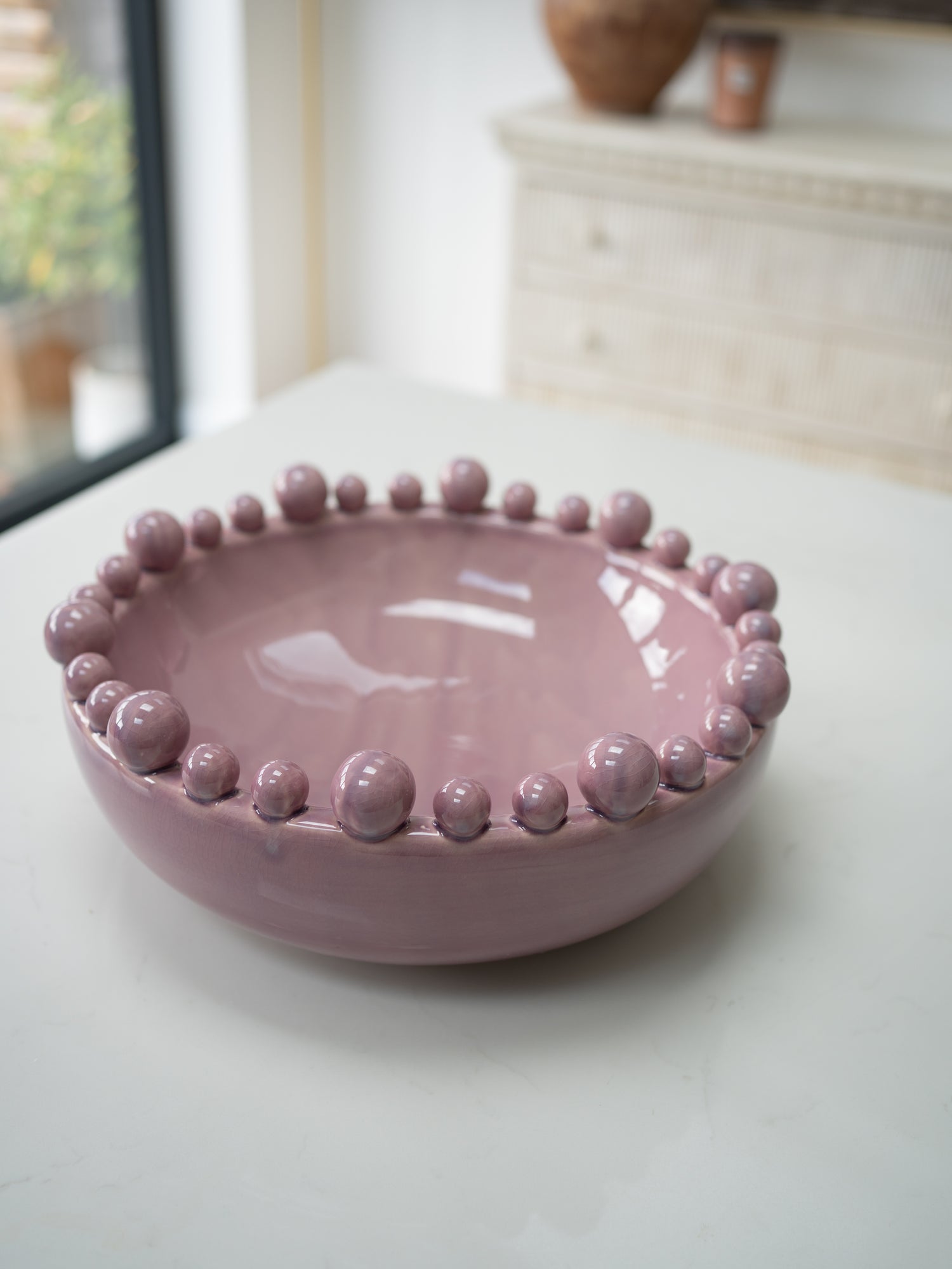 A dusty pink glazed ceramic bowl with a raised beaded rim, set on a light-colored tabletop in a softly blurred indoor setting.