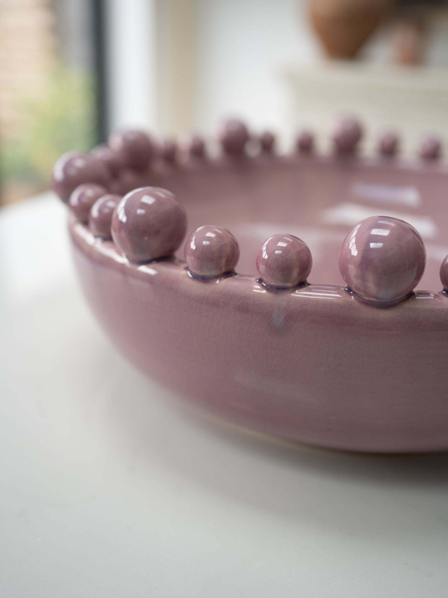 A close-up of a dusty lilac ceramic bowl with a raised beaded rim, resting on a white surface with a softly blurred background.