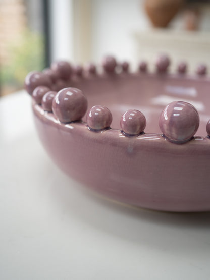 A close-up of a dusty lilac ceramic bowl with a raised beaded rim, resting on a white surface with a softly blurred background.