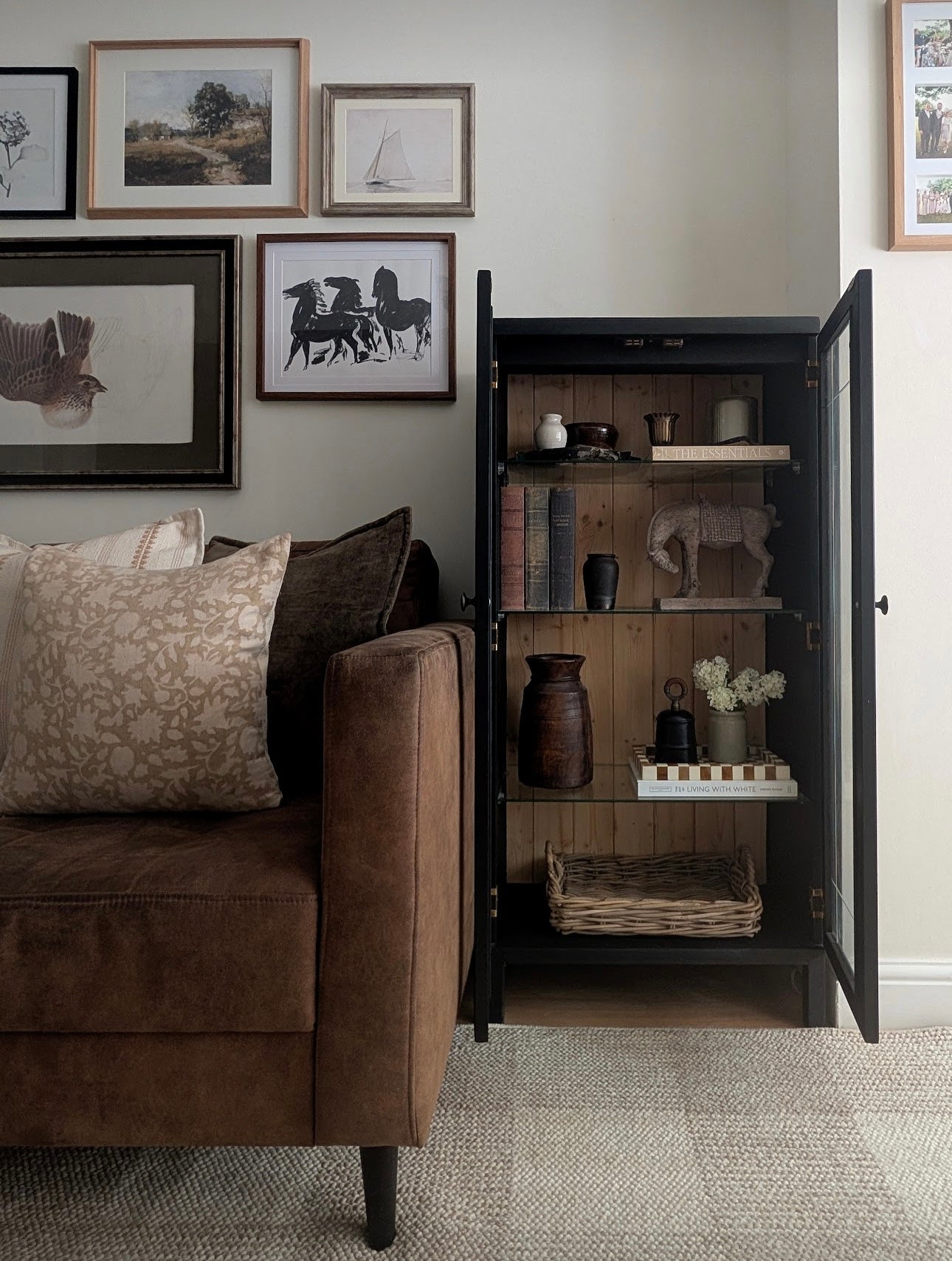 A tall black cabinet with glass doors and wooden shelves stands open beside a brown sofa in a styled living room. The cabinet is displayed with books, vases, a small elephant figurine, and other decor items inside.
