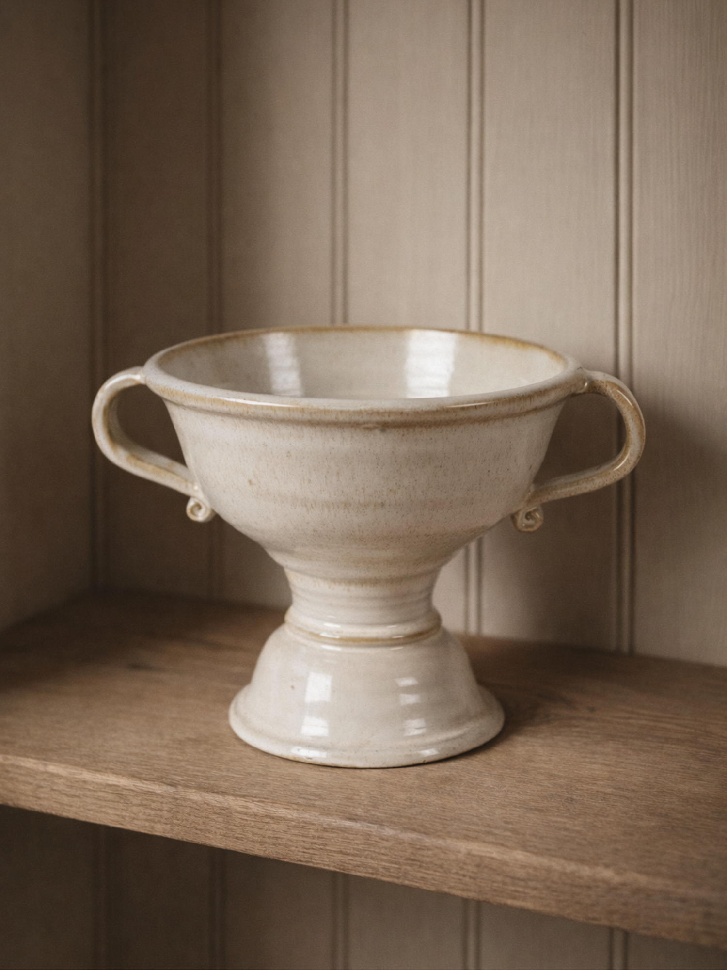 White ceramic footed vase with handles on a wooden shelf against a beige paneled wall.