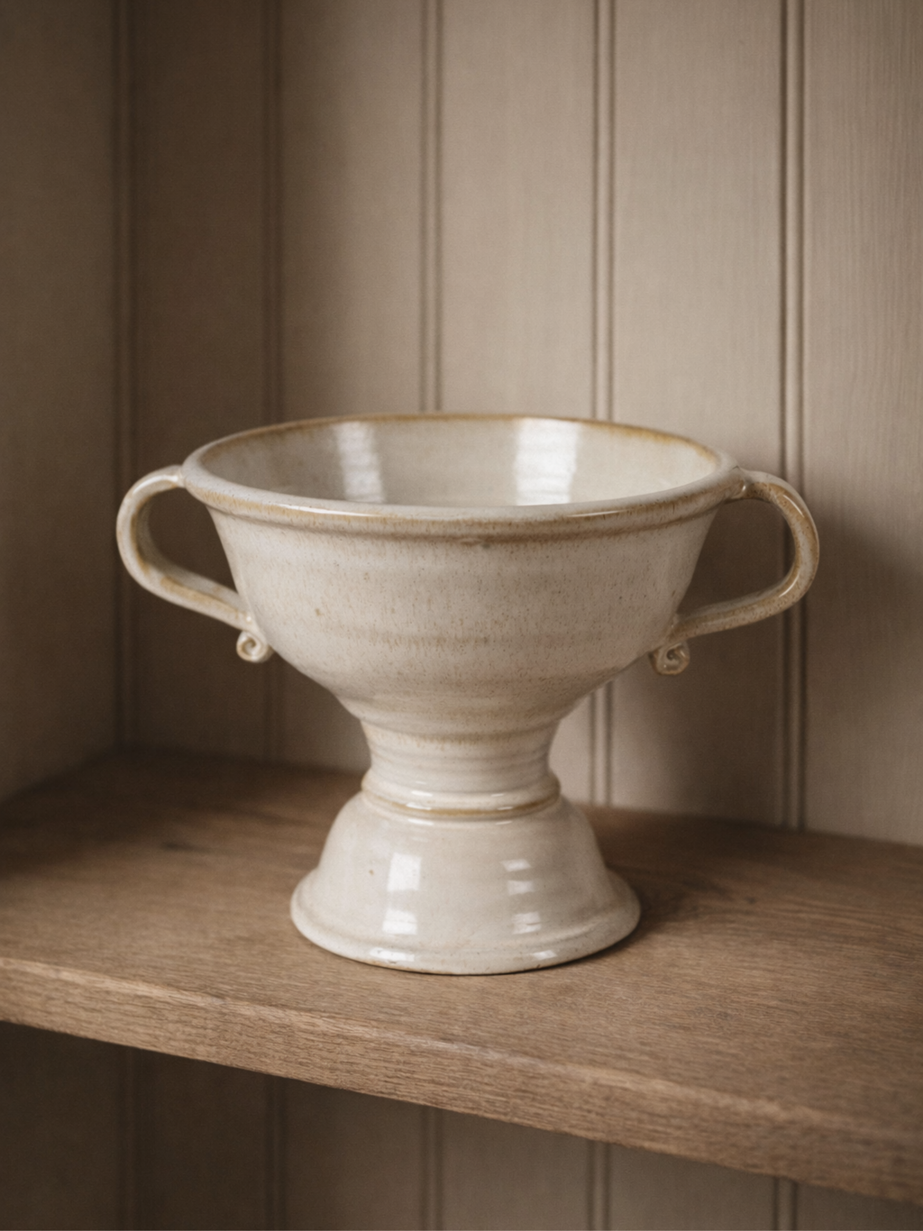 White ceramic footed vase with handles on a wooden shelf against a beige paneled wall.