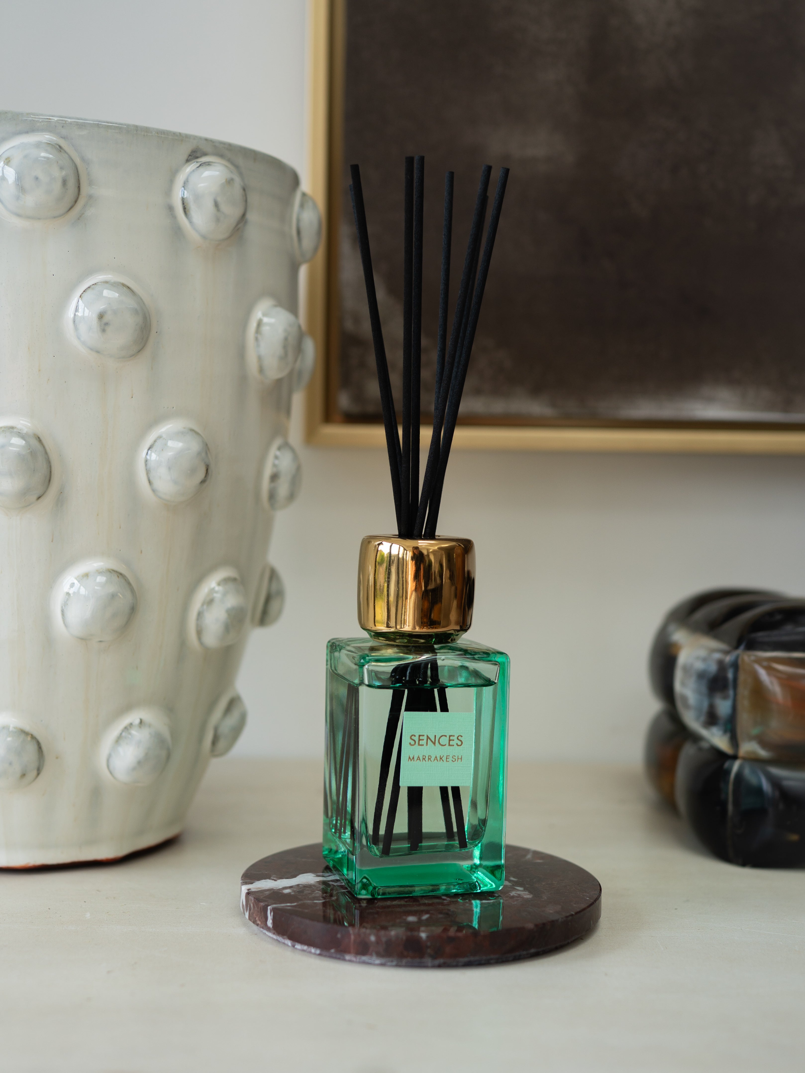 A SENCES reed diffuser bottle with a green glass body, gold cap, and black reeds, resting on a round dark coaster on a tabletop.