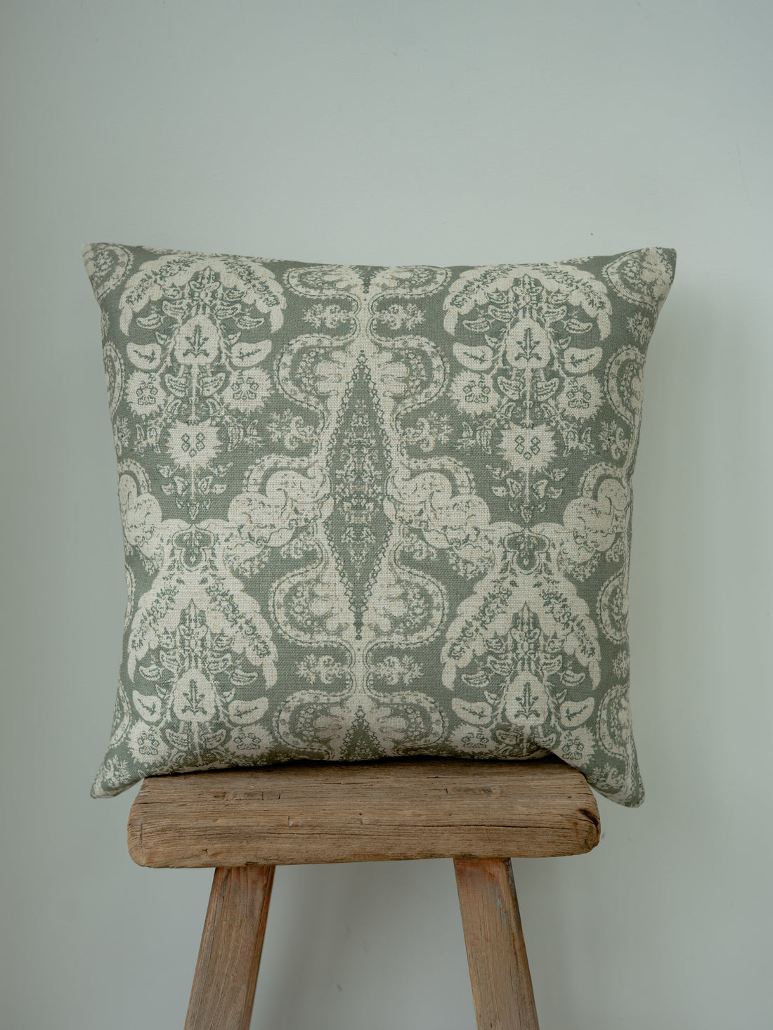 A square throw cushion with a muted sage green and cream damask pattern, resting on a rustic wooden stool against a plain light gray wall.