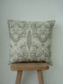 A square throw cushion with a muted sage green and cream damask pattern, resting on a rustic wooden stool against a plain light gray wall.
