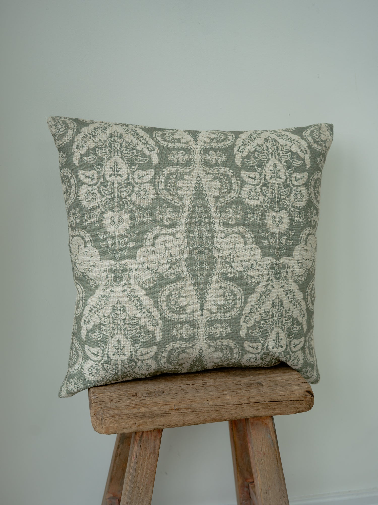 A square decorative throw cushion with a muted sage green and cream damask pattern, displayed on a rustic wooden stool against a plain light gray wall.
