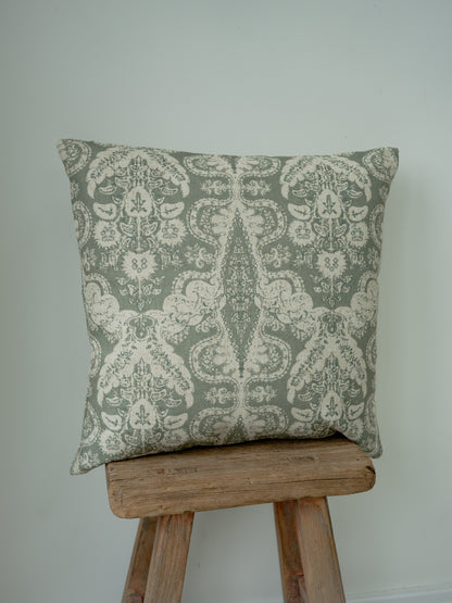 A square decorative throw cushion with a muted sage green and cream damask pattern, displayed on a rustic wooden stool against a plain light gray wall.
