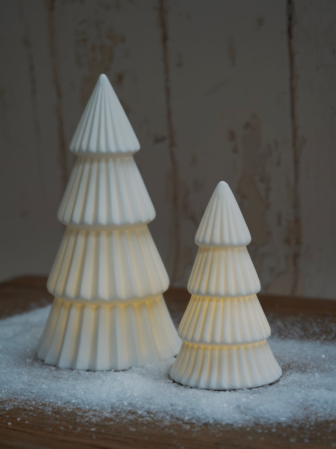 Two white ceramic Christmas trees on a wooden surface with a snowy background.