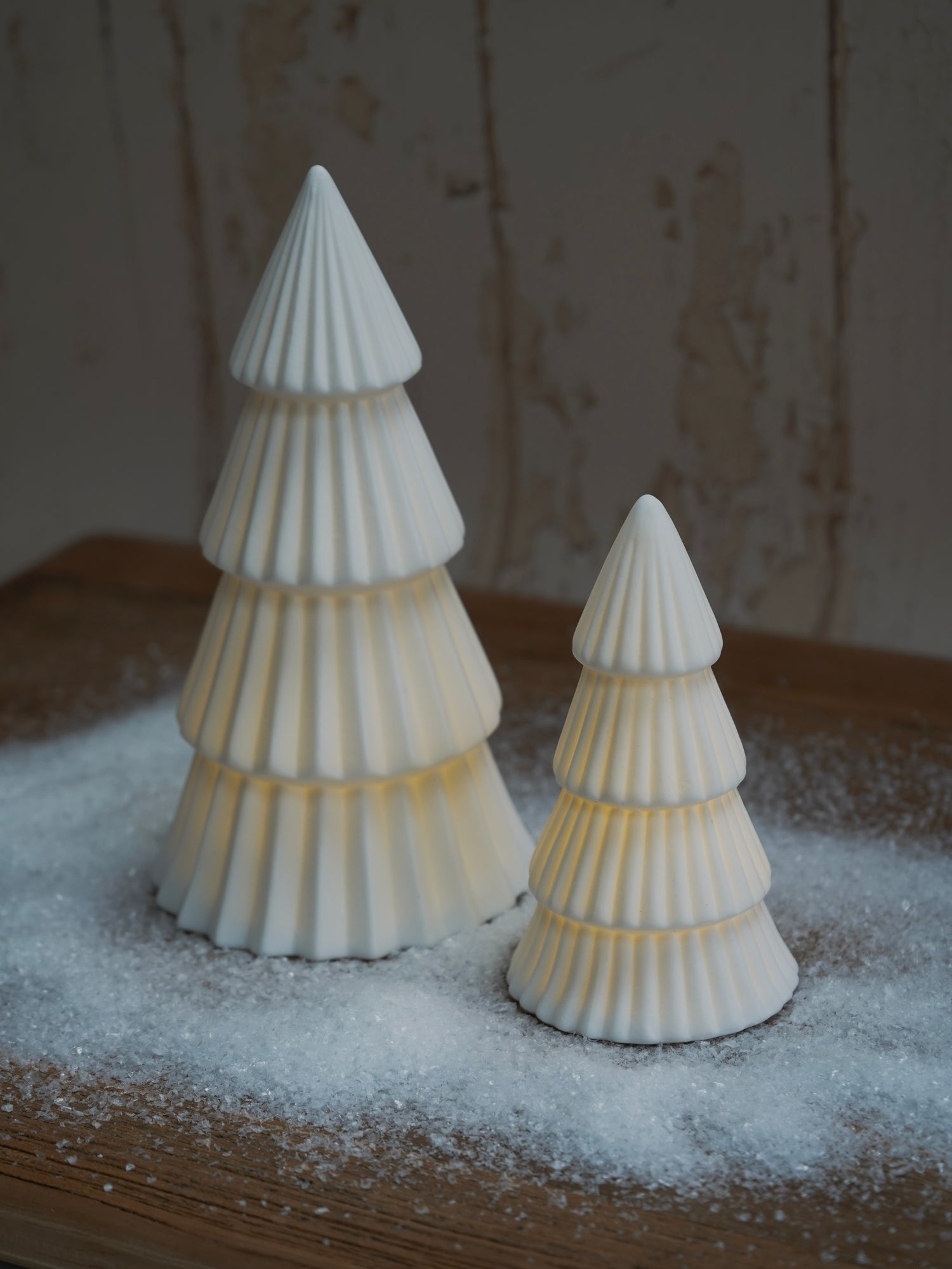 Two white ceramic Christmas trees on a wooden surface with a snowy effect.