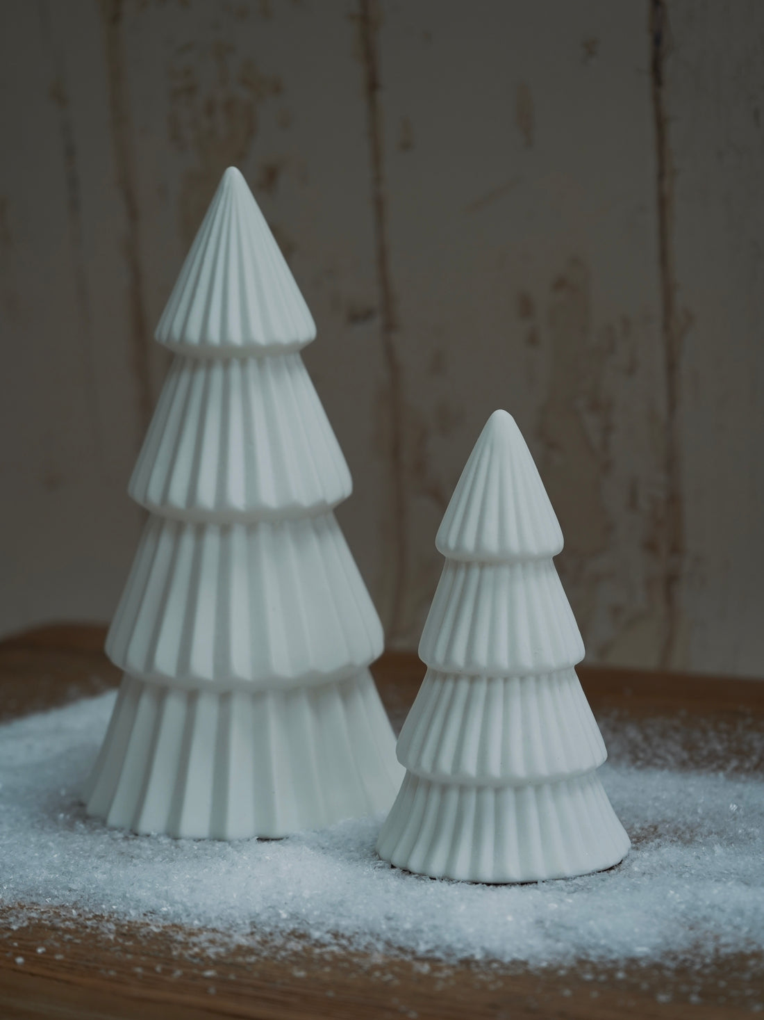 Two white ceramic Christmas trees on a wooden surface with a snowy background