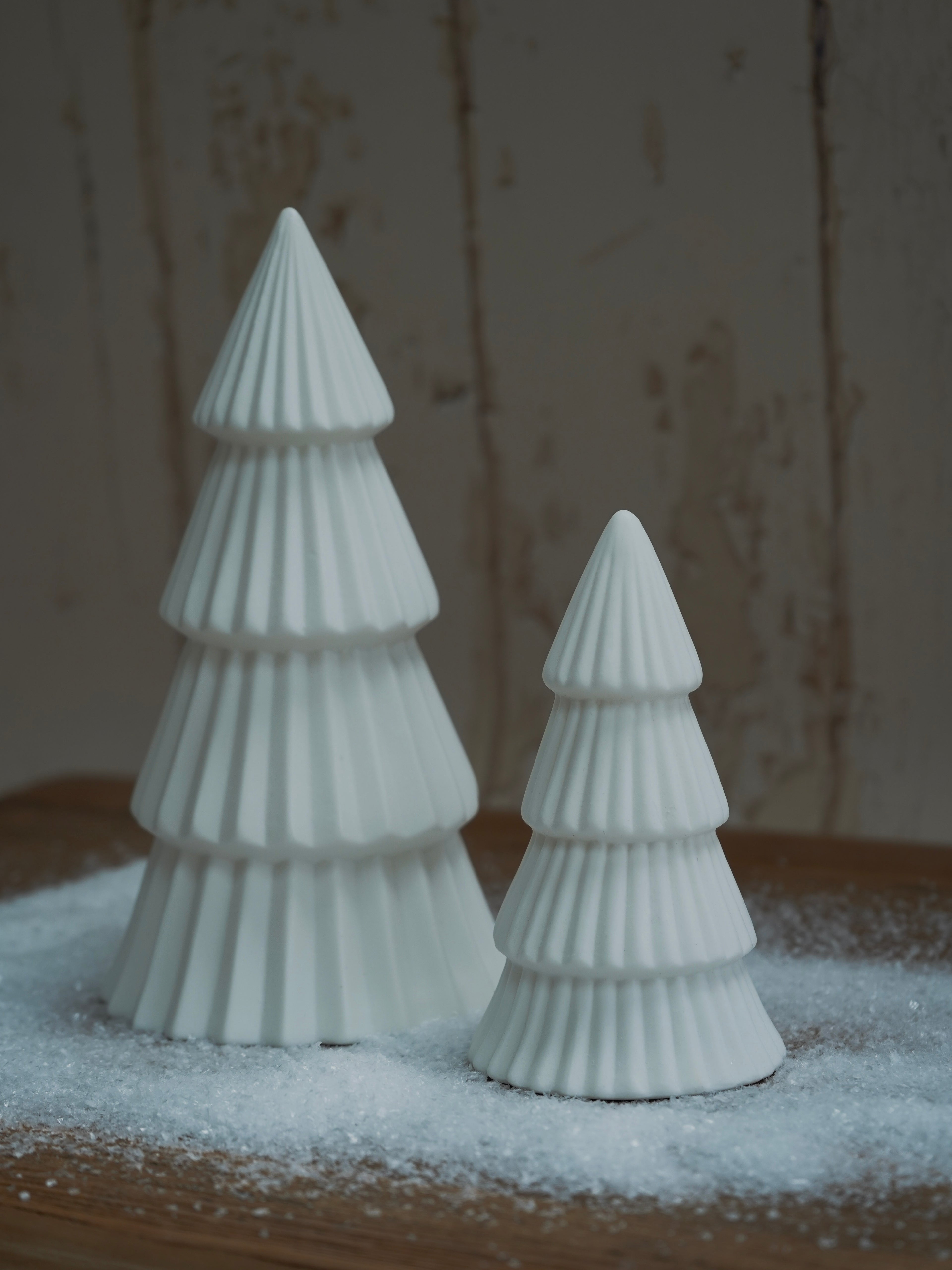 Two white ceramic Christmas trees on a wooden surface with a snowy background
