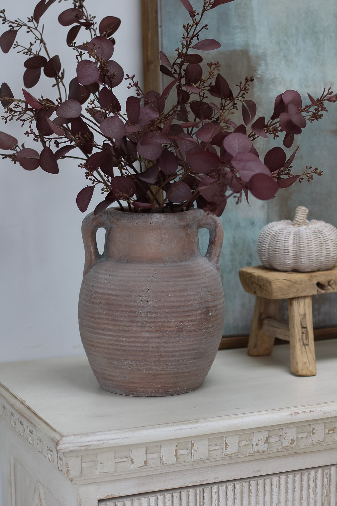 Decorative vase with purple foliage on a white surface with a wooden stool and pumpkin in the background.