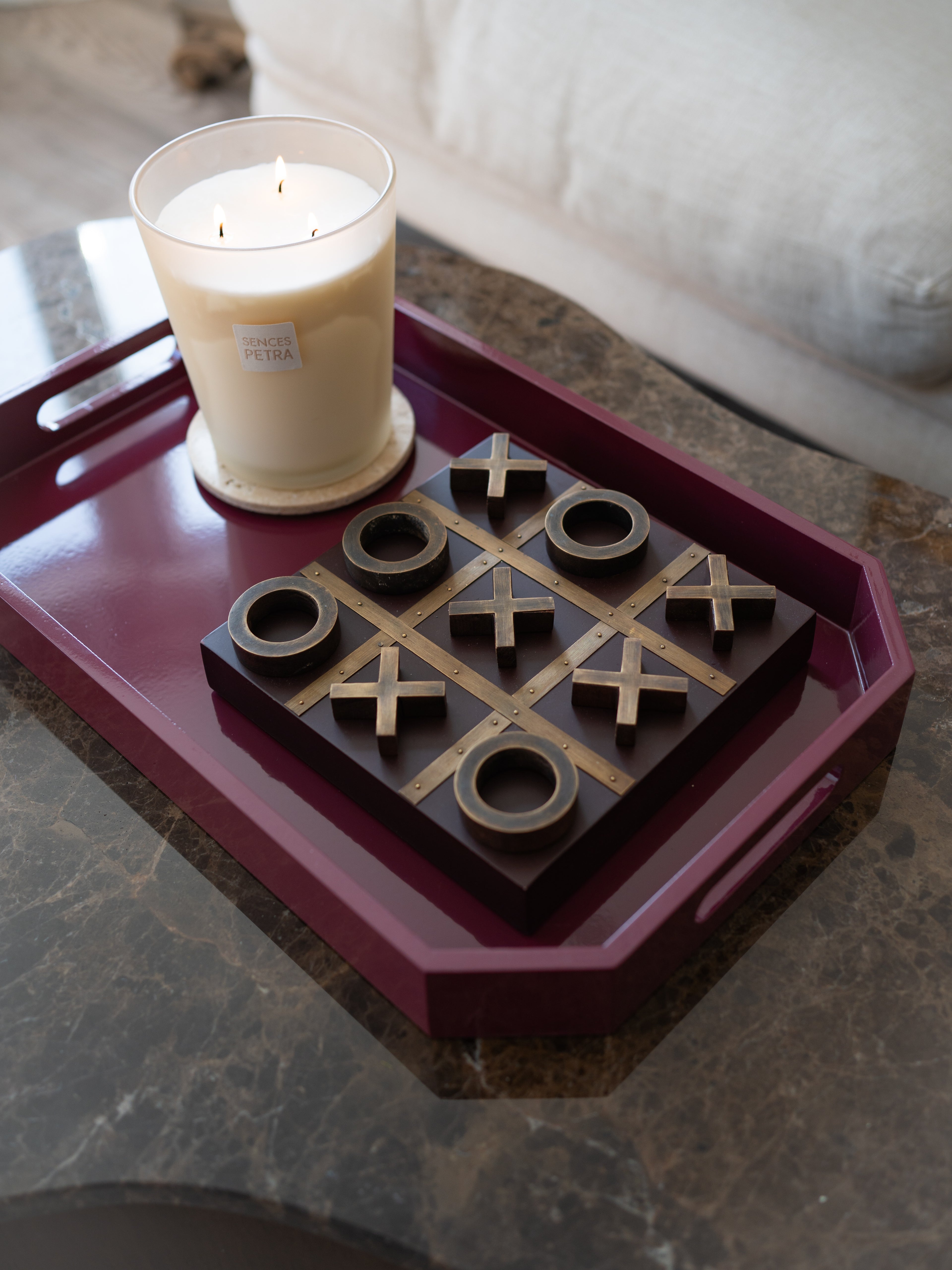 A dark wood tic-tac-toe game board with metallic gold grid lines and circular pieces, styled on a deep burgundy tray next to a lit white candle on a marble table.
