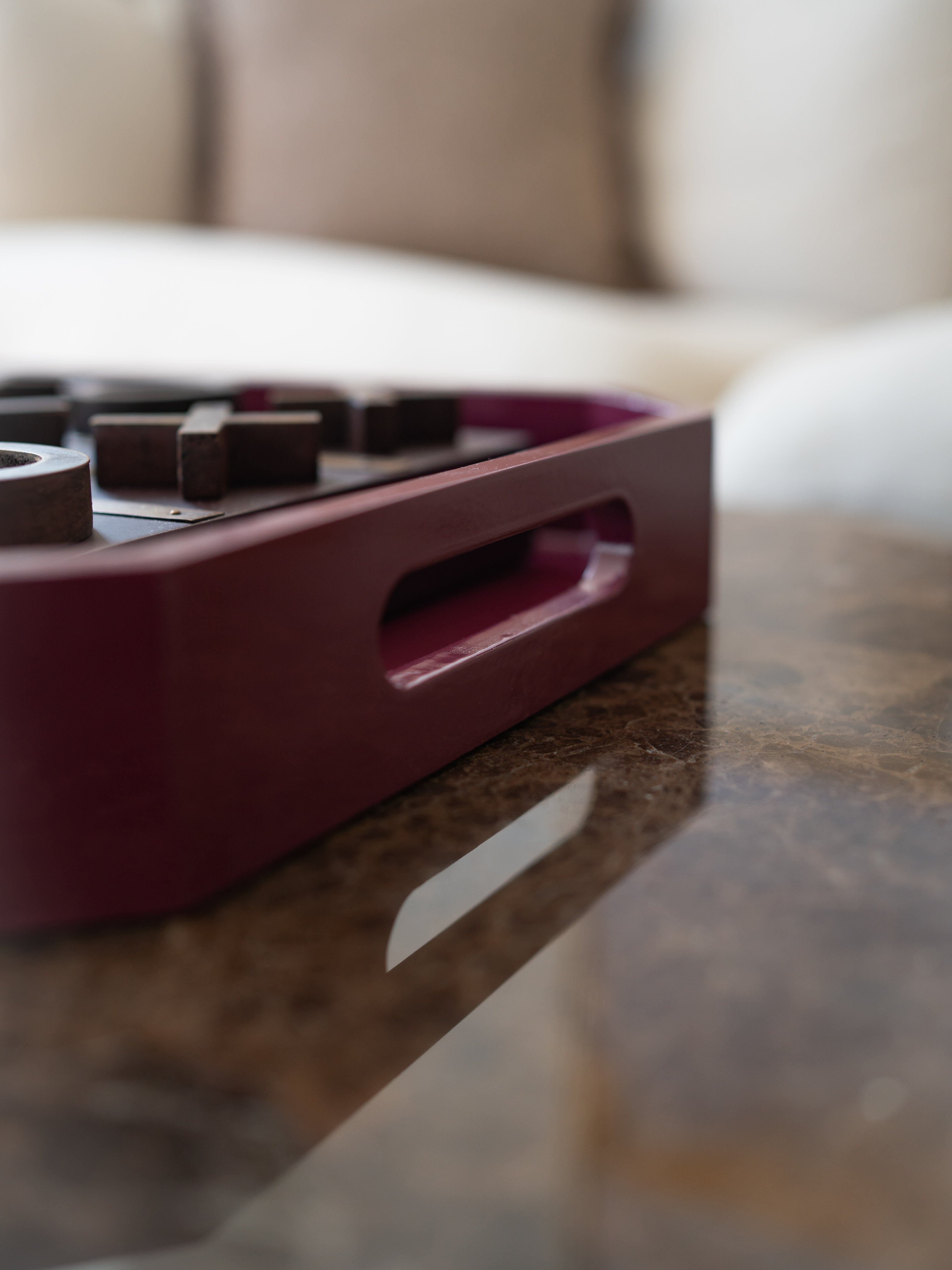 A close-up shot of a dark burgundy wooden game tray containing dark wooden pieces, resting on a polished brown marble table with a blurred living room background.