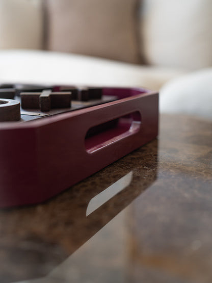 A close-up shot of a dark burgundy wooden game tray containing dark wooden pieces, resting on a polished brown marble table with a blurred living room background.