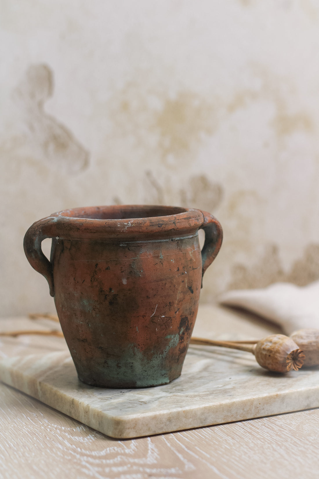 A small terracotta vintage olive pot with handles, showing signs of aging and use, placed on a marble surface.