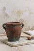 A small terracotta vintage olive pot with handles, showing signs of aging and use, placed on a marble surface.