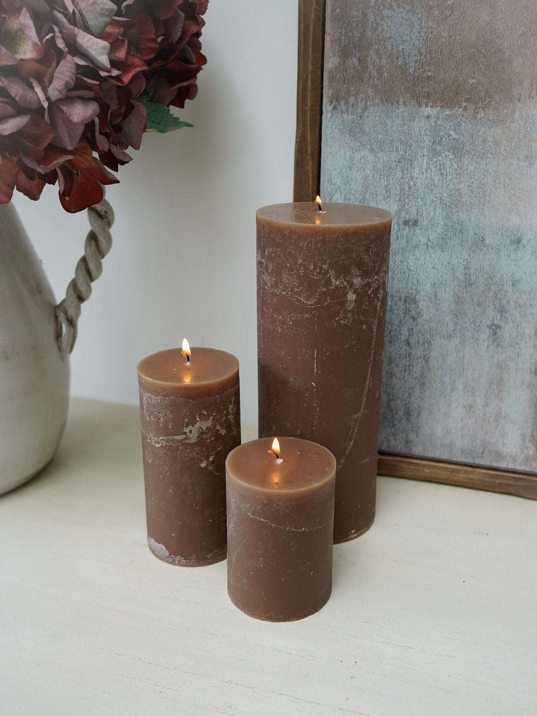 Three lit mocha pillar candles of different sizes on a white surface with a decorative vase and mirror in the background.