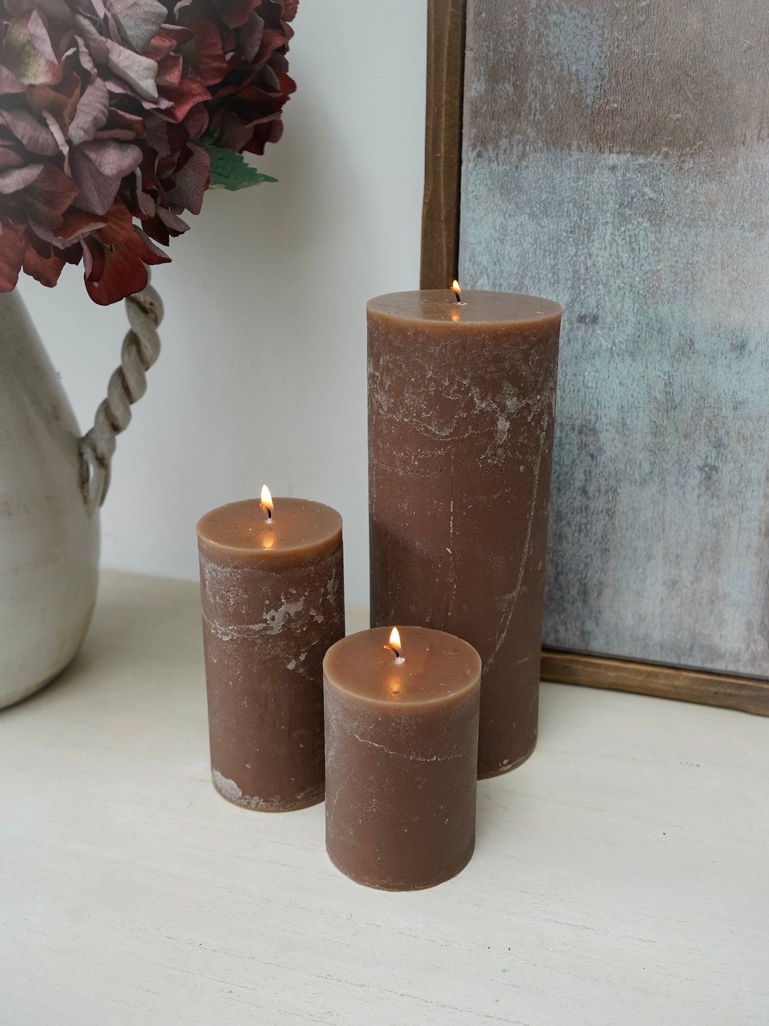 Three lit mocha pillar candles of different sizes on a white surface with a decorative vase and mirror in the background.