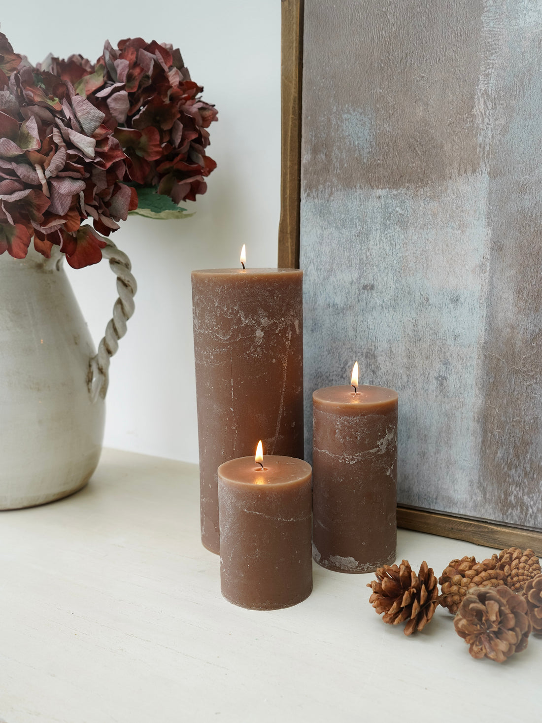 Three lit mocha toned pillar candles on a surface with a vase of flowers and decorative items.
