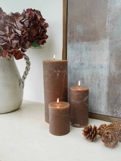Three lit mocha toned pillar candles on a surface with a vase of flowers and decorative items.