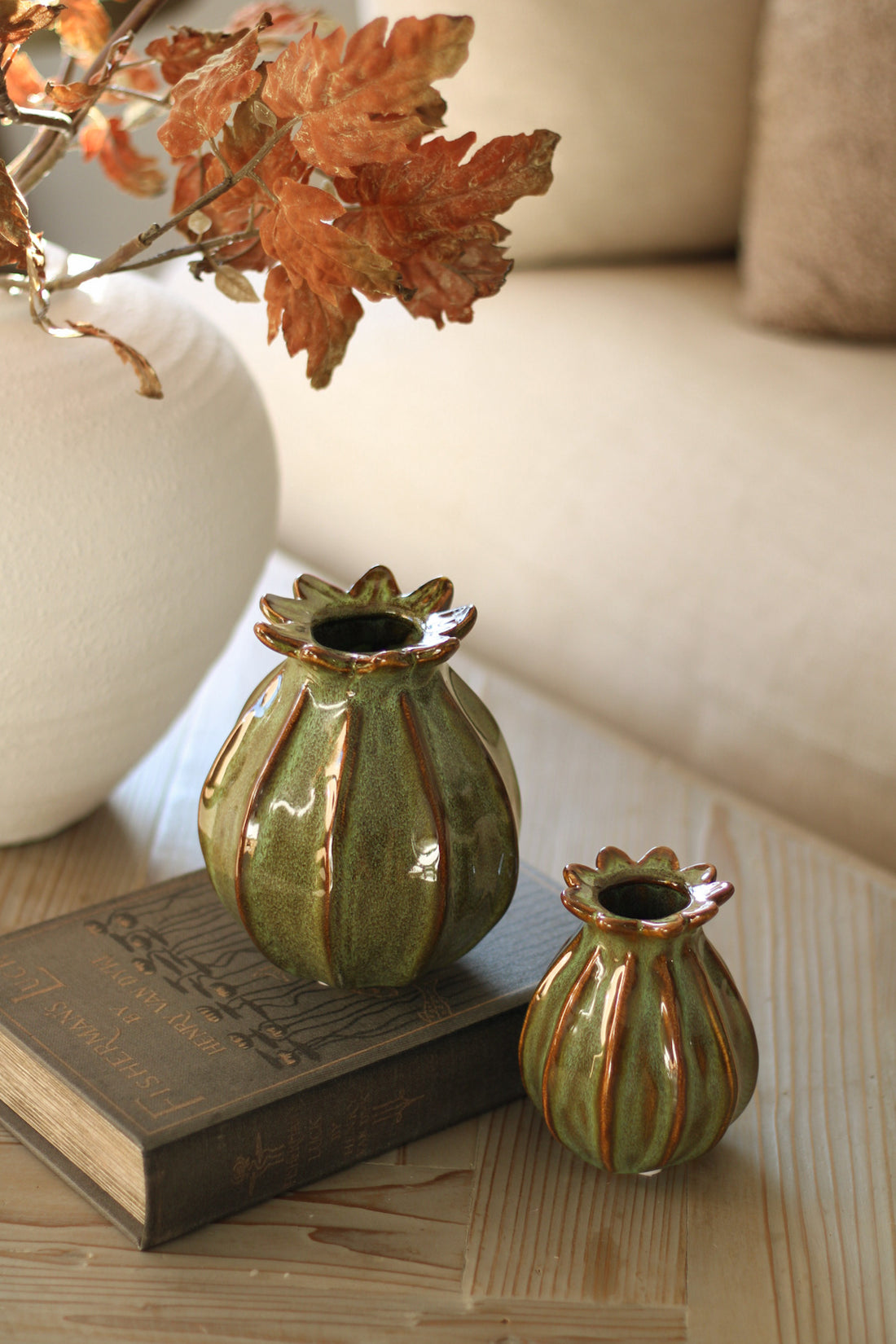 Two green ceramic poppy head vases with brown stripes on a wooden surface with a book and decorative leaves.