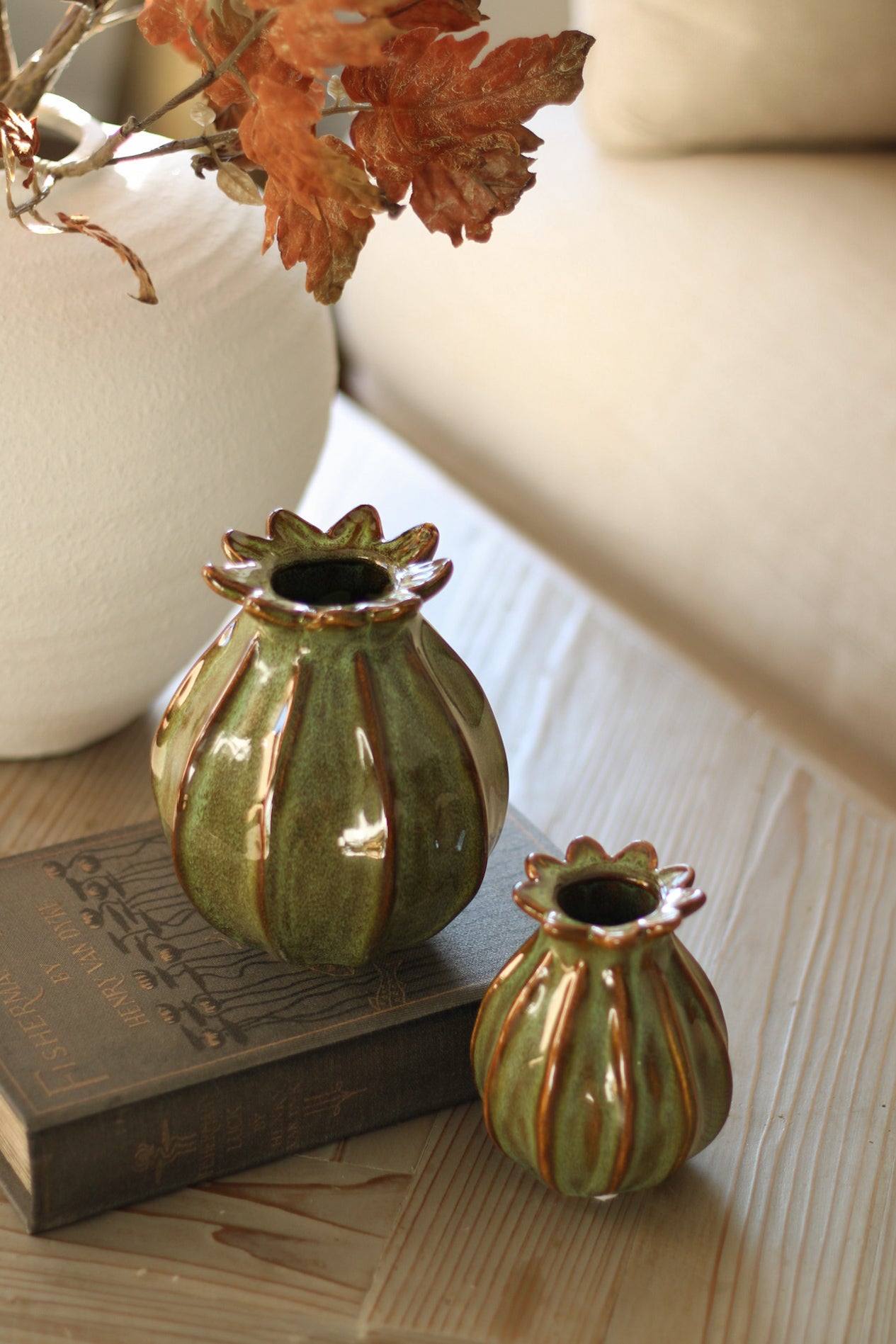 Two green ceramic poppy head vases with gold accents on a wooden surface with a book and leaves.